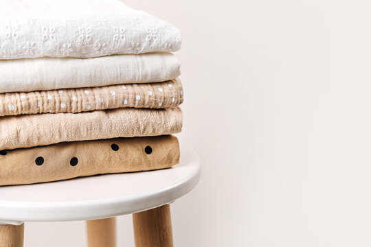 Stack of baby clothes. Cotton clothes and muslin swaddle blanket in white and beige colors. Clean freshly laundered, neatly folded kids clothes on table.