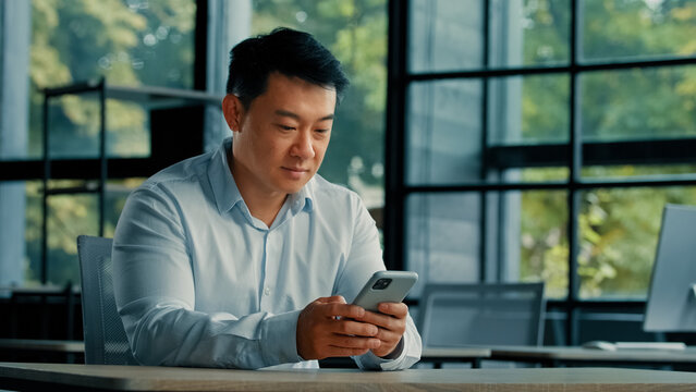 Male portrait asian chinese man entrepreneur employer work in office look at modern gadget phone mobile screen search information online use smartphone apps technology 5g wi-fi online communication