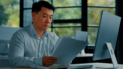 Fototapeta premium Professional asian businessman korean japanese 40s man lawyer office worker reading financial documents invoice contract budget documentation searching business data at computer paying bills online