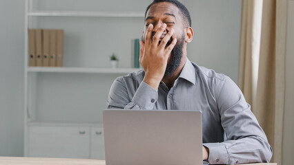 Tired exhausted ethnic African American male worker yawning sitting at table in office law energy burnout insomnia. Overworked businessman professional man feel fatigue at boring work yawn need sleep