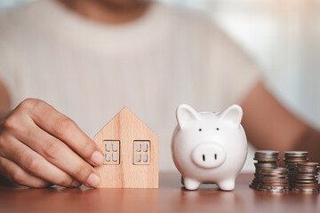 Asian women's hand holding a wooden house model with a saving piggy bank and a pile of coins and money on the wooden table for business, finance, saving money and property investment concept.