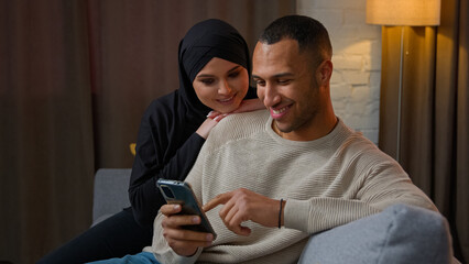 Family couple multiethnic interracial family African American man chatting on mobile phone muslim woman wife cuddle to husband use cellphone shopping online together watch funny internet video at home