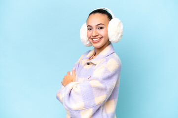Young Arab woman wearing winter muffs isolated on blue background with arms crossed and looking...