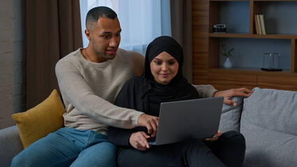 Married couple sit on sofa use laptop device choosing new apartment on internet website multiracial family shocked amazed diverse man woman search information by computer read good news talking indoor