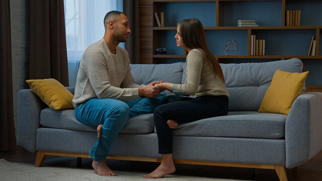 Multiracial couple in love married family man husband talk with woman wife hold hands supporting relationship care romantic intimacy date sit on sofa in living room girl guy talking tender relations