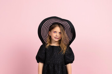 Portrait of a Charming little girl in black stylish summer dress, sun hat and a slight makeup. Isolated on pink background.