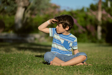 Fototapeta premium child sunny day, sitting on grass and covering his eyes from sunlight to get a better view of something intriguing. sun safety and importance of protecting their eyes from harmful UV rays