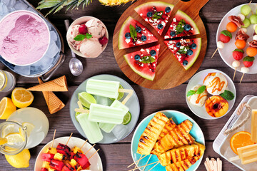 Refreshing summer food table scene. Assorted grilled fruits, ice cream and ice pops. Above view on a dark wood background.