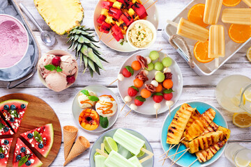 Refreshing summer food table scene. Assortment of grilled fruits, ice cream and ice pops. Top down view on a white wood background.