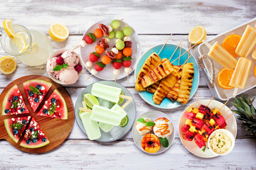 Refreshing summer food table scene. Assorted grilled fruits, ice cream and ice pops. Top view on a white wood background.