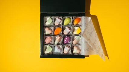 Close up of homemade multicolored chocolate bonbons in a gift box. Assortment of hand painted candies. Flat lay. Macro shot of chocolatier products on the yellow background.