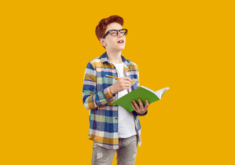 Studio shot of pensive school child. Student boy in casual plaid shirt and eyeglasses standing isolated on yellow background, doing homework, holding pencil and notebook, looking away and thinking