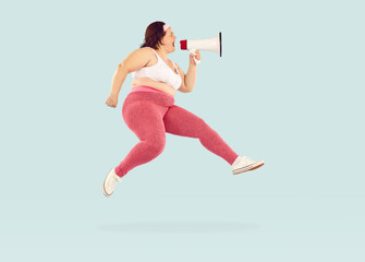 Fat overweight woman wearing sportswear holding mouthpiece on studio blue background and jumping. Plump girl shouting and screaming announcement in megaphone. Advertising and body positive concept.