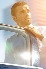 A middle-aged man stands next to a car in the city in the morning, the sun is rising, the rays are bright.