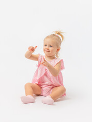 A 1-2-year-old Caucasian girl in a pink dress is sitting on the floor looking and pointing with her fingers on a white background. Copy space.