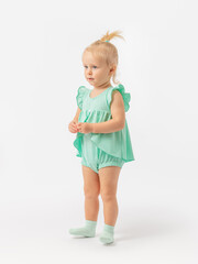 A 2-year-old baby in a green dress and socks stands on her heels on a white background. Exercise, exercise.