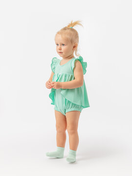 A 2-year-old baby in a green dress and socks stands on her heels on a white background. Exercise, exercise.