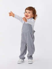 A 2-year-old toddler with curly hair smiles broadly, laughs and stretches his arms up in a gray jumpsuit and a white turtleneck and full-length sneakers on a white background.