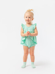 A 2-year-old toddler girl stands smiling and looks expressively at a white background with interest in a green bodysuit dress and socks.