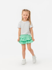A 5-year-old girl with long hair looks expressively at the camera in a T-shirt and holds on to a skirt-shorts, bending her knee in sneakers on a white background. photo