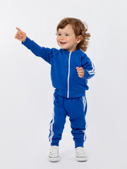 A cheerful little boy of 1-2 years old stands, smiles and points his finger up in a blue tracksuit and sneakers on a white background.
