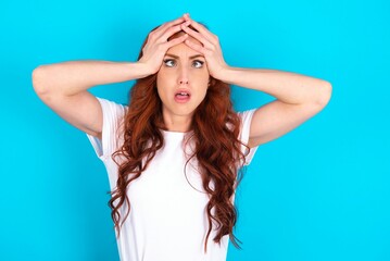 Horrible, stress, shock. Portrait emotional crazy young redhead woman wearing white T-shirt over blue background clasping head in hands. Emotions, facial expression concept.