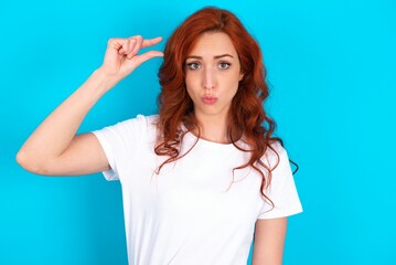Fototapeta premium Shocked young redhead woman wearing white T-shirt over blue background shows something little with hands, demonstrates size, opens mouth from surprise. Measurement concept.