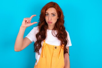 Young redhead woman wearing orange overall over blue background purses lip and gestures with hand, shows something very little.