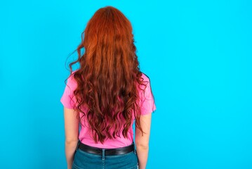 The back view of a young redhead woman wearing pink T-shirt over blue background . Studio Shoot.
