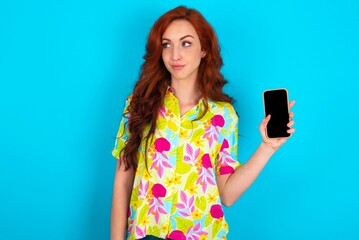 Young redhead woman wearing colorful shirt over blue background holds new mobile phone and looks mysterious aside shows blank display of modern cellular