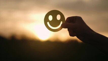 Selective focus of male hand holding yellow happy smile face on background amazing sunset or sunrise, good feedback rating and positive customer review, satisfaction survey, mental health assessment