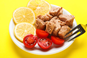 Plate with delicious canned tuna, fresh lemon, tomatoes and fork on yellow background, closeup