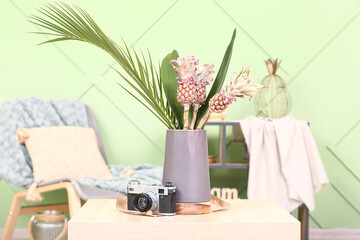 Vase with baby pineapples, palm leaves and photo camera on table in living room