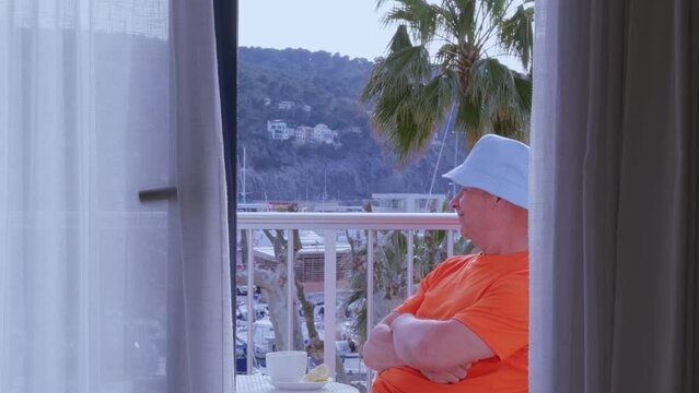 mature man in orange t-shirt, senior of 60 years old sitting on balcony, enjoys view outskirts palm trees, yachts, resort town port Soller on island Mallorca, enjoying journey, vacation, resort place