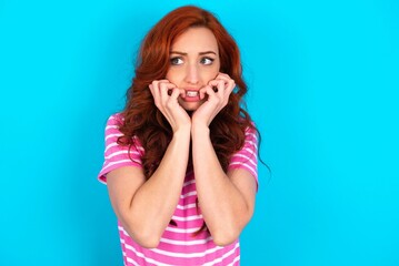 Terrified young redhead woman wearing striped T-shirt over blue background looks empty space home alone moonless night