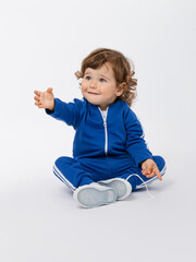 Curly-haired toddler 1-2 years old stretches out his hand and asks for help to get up, looks expressively in a blue tracksuit and sneakers on a white background