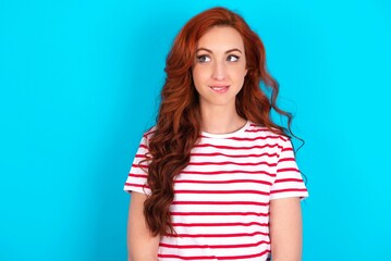 Amazed young redhead woman wearing striped T-shirt over blue background bitting lip and looking tricky to empty space.