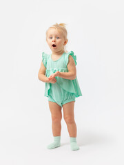 Surprise, indignation, question. A 2-year-old girl stands with folded arms on a white background in a green bodysuit dress with an expressive look.