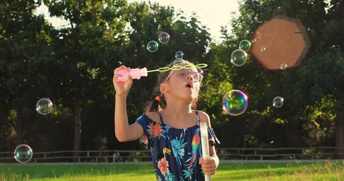 Cute little girl blowing soap bubbles, having fun outdoors on backyard on summer warm day. Kid playing on holiday at countryside. Happy children day. Cheerful and carefree childhood.