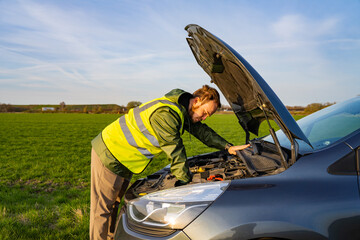 Young man in yellow reflective vest examining his car engine parked on the side of a road