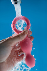 Woman holding pink anal beads under running water on blue background. Sex toy hygiene concept.