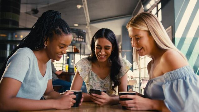Group of multi-cultural female friends meeting in restaurant or coffee shop looking at social media on mobile phone and laughing together - shot in slow motion
