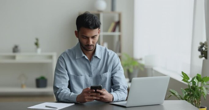 Serious Indian man sitting at desk in home office, distracted from work on computer, browsing web on smart phone, typing answer, messaging to client, lead personal chat, use modern Generative AI