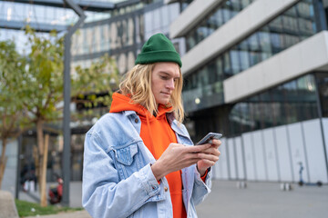 Millennial hipster guy with blond hair browsing internet on mobile phone, standing on city street,...