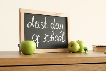 Chalkboard with text LAST DAY OF SCHOOL and apples on table near light wall