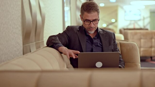 Businessman working in business lounge. Entrepreneur sitting on sofa in office lobby using laptop computer to do remote work online. Happy middle aged, mid adult, mature age man smiling.
