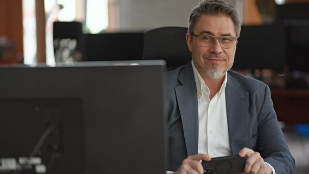 Business portrait - confident businessman sitting at desk working in office, using phone. Happy mid adult man in shirt and jacket, smiling. Bearded, glasses.