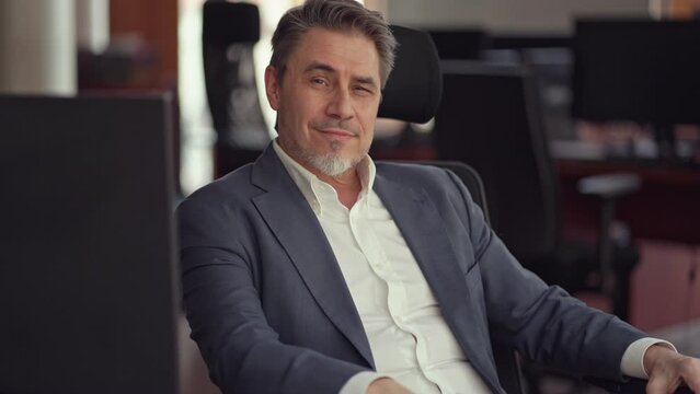 Business portrait - confident businessman sitting at desk in office. Happy mid adult man in shirt and jacket, smiling. Bearded, gray hair.