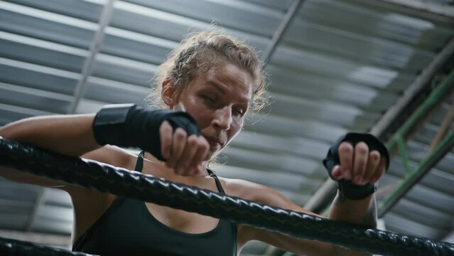 Woman boxer wet with sweat takes a deep breath leaning ring ropes in training. Portrait of tired attractive female athlete resting between fights at gym competition