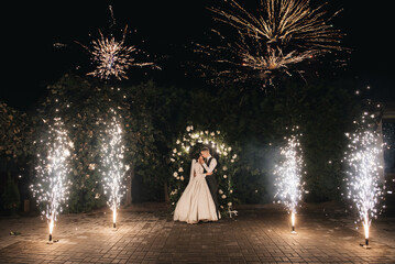 Young bride and groom stand near the wedding arch at night with lights, smoke and fireworks,...
